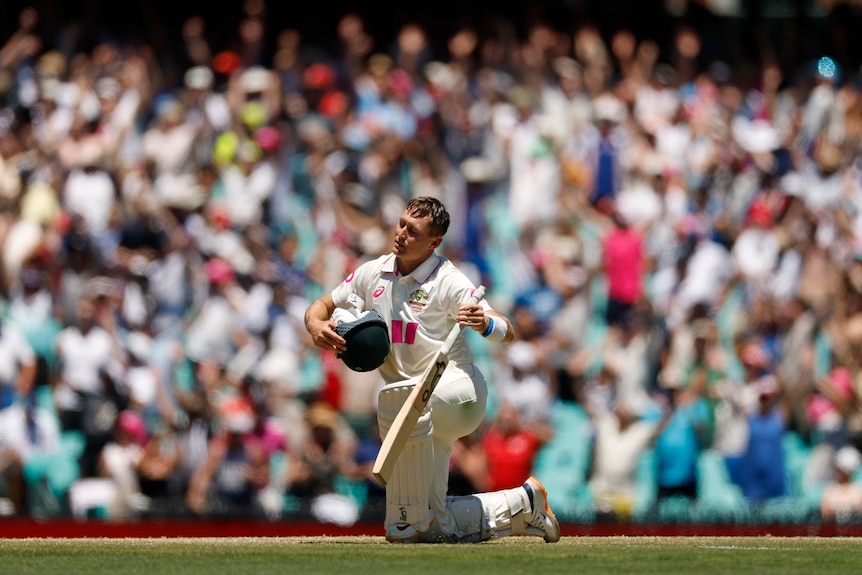 A cricket batter is on his knees in the middle of the pitch, holding bat and helmet