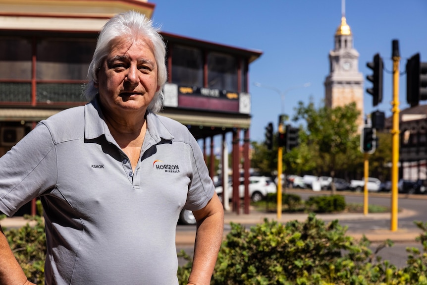 A silver-haired, older man in a polo shirt stands across the road from a pub in a historic-looking CBD.