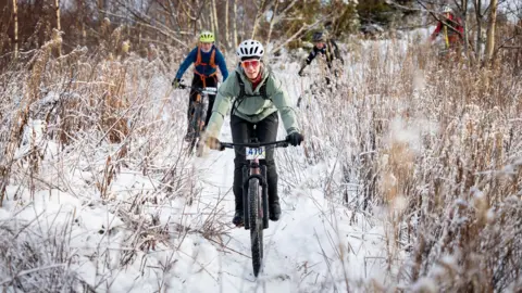 Paul Campbell A mountain biker wearing a white helmet, reflective glasses, light green jacket and black waterproof trousers rides through a snow-covered area of long grass. Behind them is another cyclist wearing a bright yellow helmet, blue jacket and black waterproof trousers.