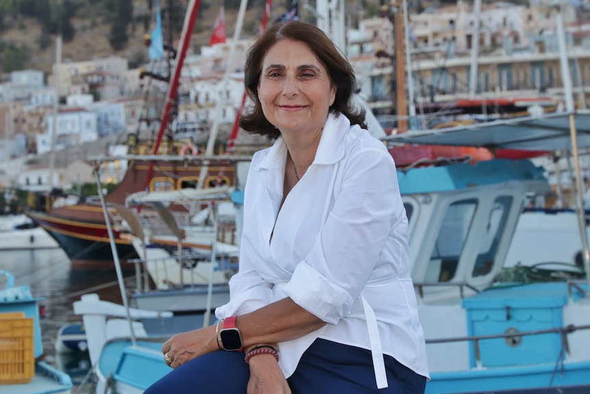 White woman, brown bob-haircut, white long-sleeve blouse, blue jeans, sitting a docking harbour in Greece.