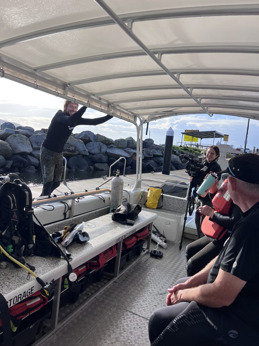 Four people in diving equipment on a boat next to a rock wall