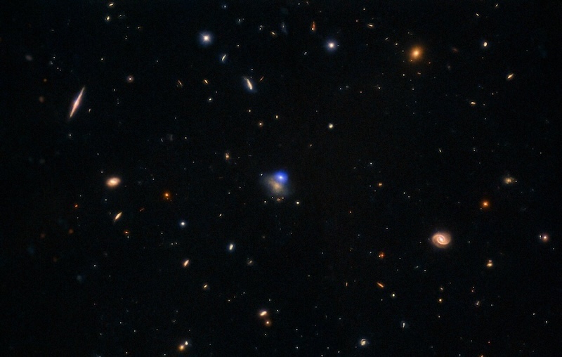 Black space, with scattered tiny galaxies, and a small bright blue spot in the middle next to one of them.