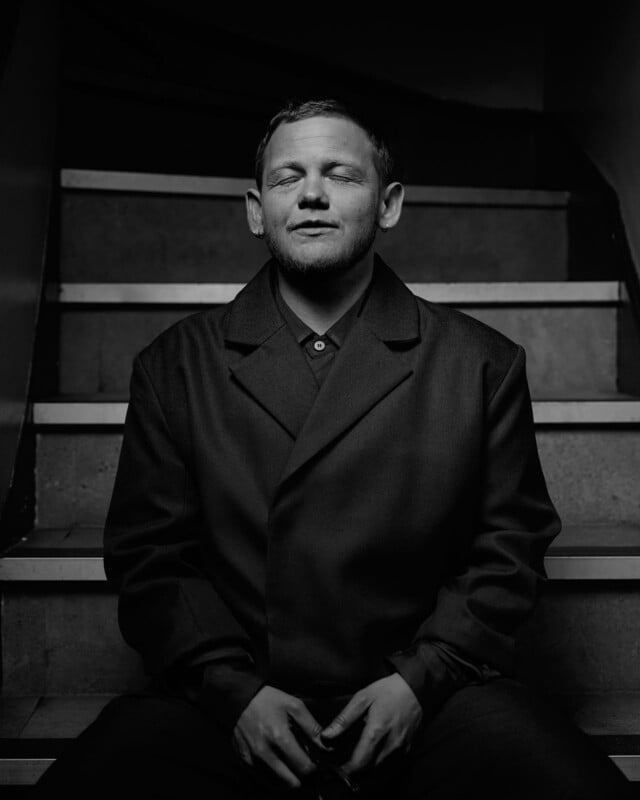 A man wearing a dark coat sits on a staircase with his eyes closed, hands resting in his lap. The photo is black and white, and the lighting highlights his relaxed expression.