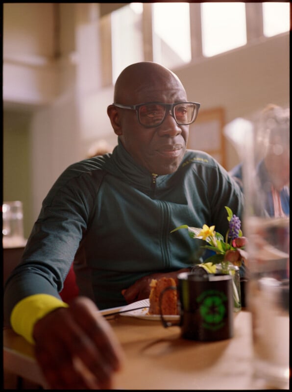An older man wearing glasses and a dark green jacket sits at a table with flowers, a mug, and a slice of cake in front of him. Sunlight streams through windows in the background.