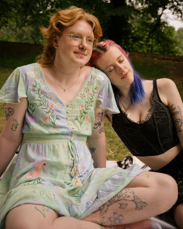 Two people sit closely together on grass. One wears a pastel dress with floral patterns and glasses, smiling. The other, with blue and pink hair, rests their head on the first person's shoulder, eyes closed, both have visible tattoos.