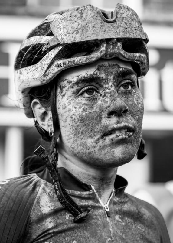 A cyclist with a helmet and goggles looks upward, their face and clothing covered in mud, suggesting participation in a challenging race. The photo is in black and white.
