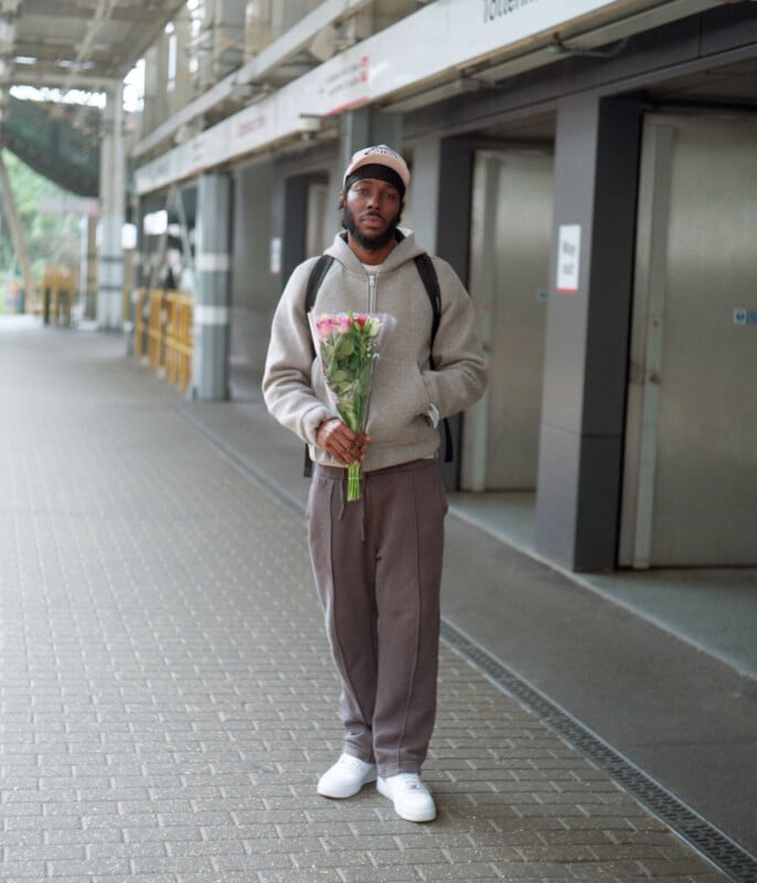 A man wearing a gray hoodie, dark pants, and white sneakers stands alone on a paved walkway holding a bouquet of pink roses. He has a backpack and is in front of a building with metal doors and overhead signage.