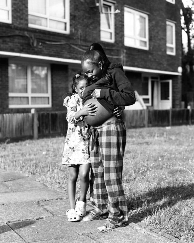 A young girl hugs a pregnant woman outdoors in front of an apartment building. The woman smiles, holding her belly, while the girl in a floral dress and sandals looks up at her affectionately.