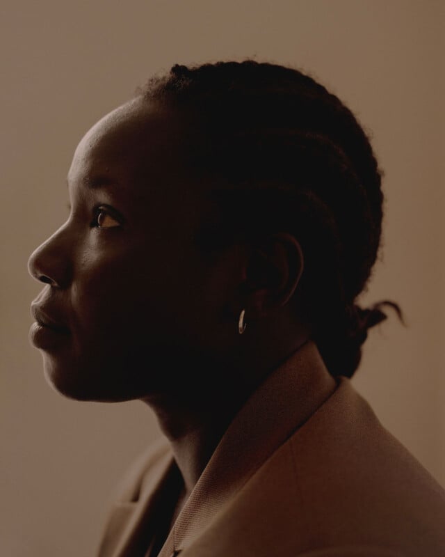 A person with braided hair and wearing a tan jacket is shown in profile against a plain, softly lit background. They have a calm expression and are wearing a small hoop earring.
