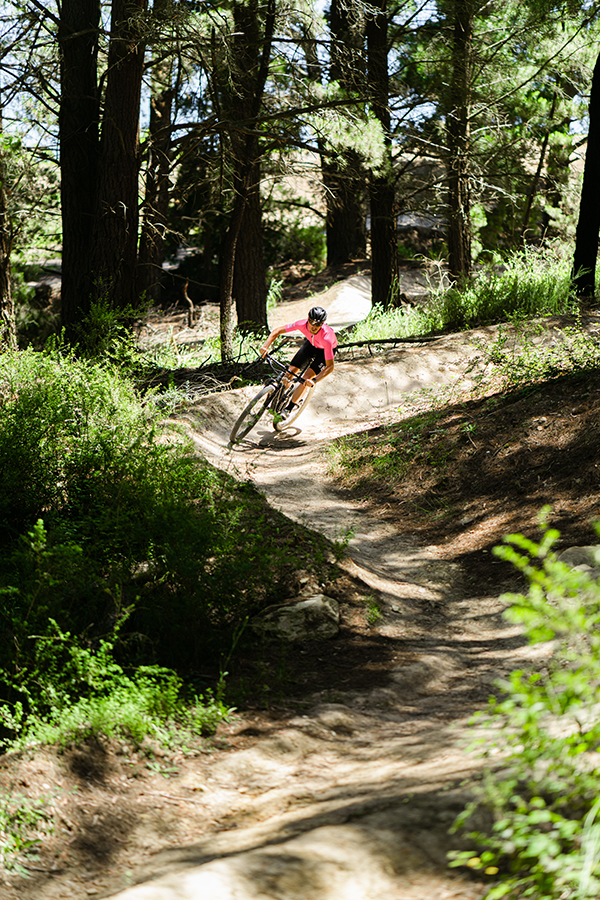 A person mountain biking through a shade bush Omeo trail, dressed in a bright pink top and dark shorts.