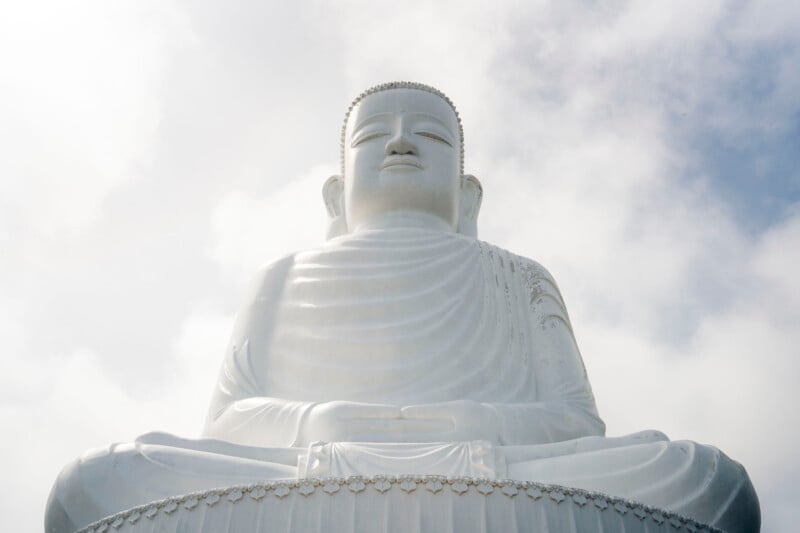 A large white Buddha statue sits in meditation against a cloudy sky, viewed from a low angle. The serene figure has detailed robes and a peaceful expression.