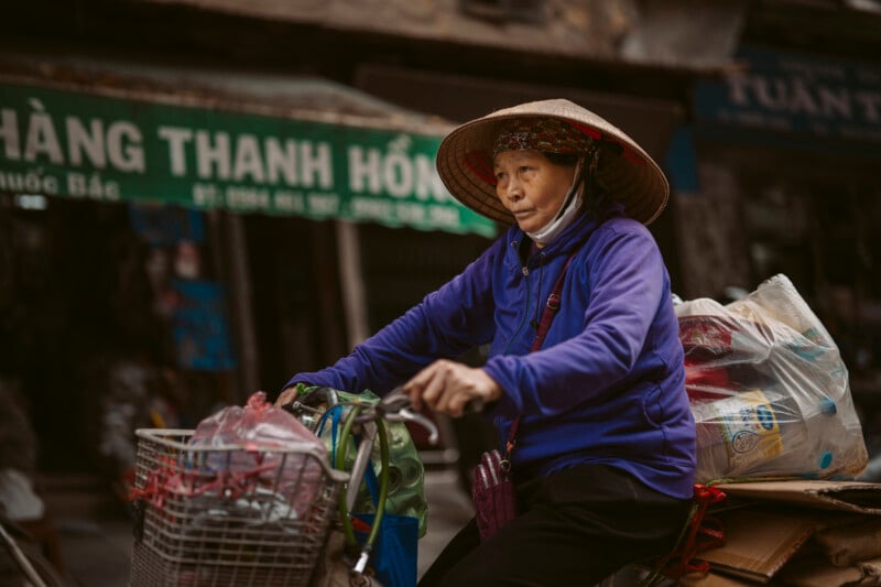 An elderly woman wearing a conical hat and blue jacket rides a bicycle loaded with bags and packages along a street, with a green shop sign visible in the background.