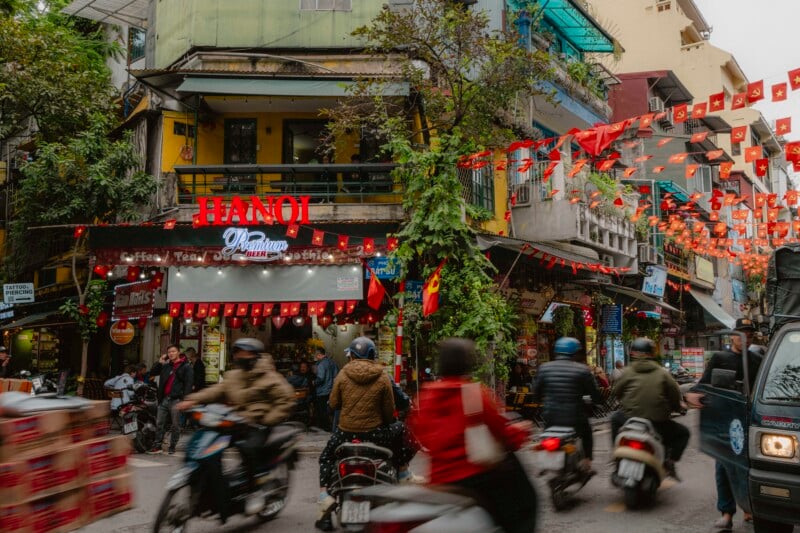 A busy street scene in Hanoi, Vietnam, with people riding motorbikes, pedestrians walking, and buildings decorated with red flags and banners. The storefront sign reads "Hanoi Premium" amidst lush greenery.