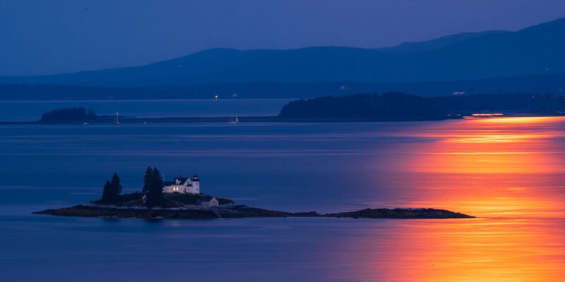 A small island with a white house and trees sits in calm blue water at dusk, illuminated by a warm, golden reflection of the sunset across the surface. Mountains are visible in the distance under the twilight sky.