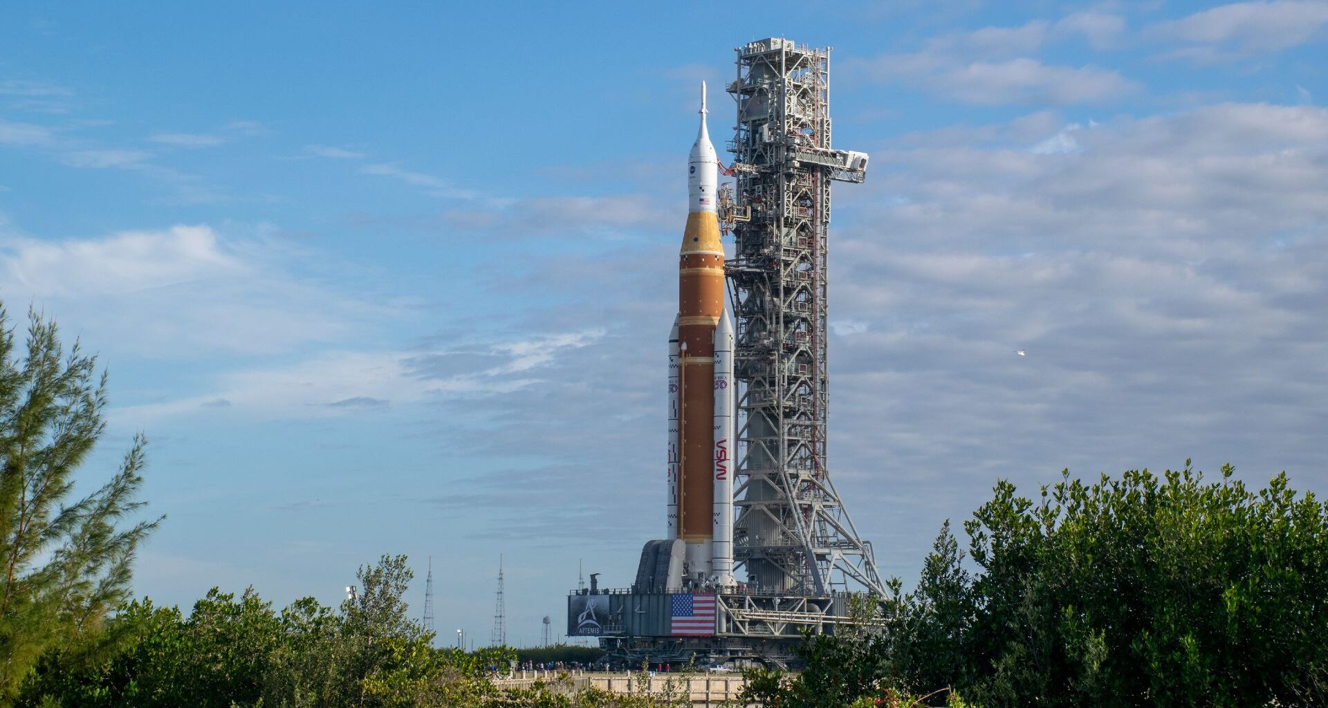 An orange rocket held up by scaffolding sits behind large green bushes under a blue sky.