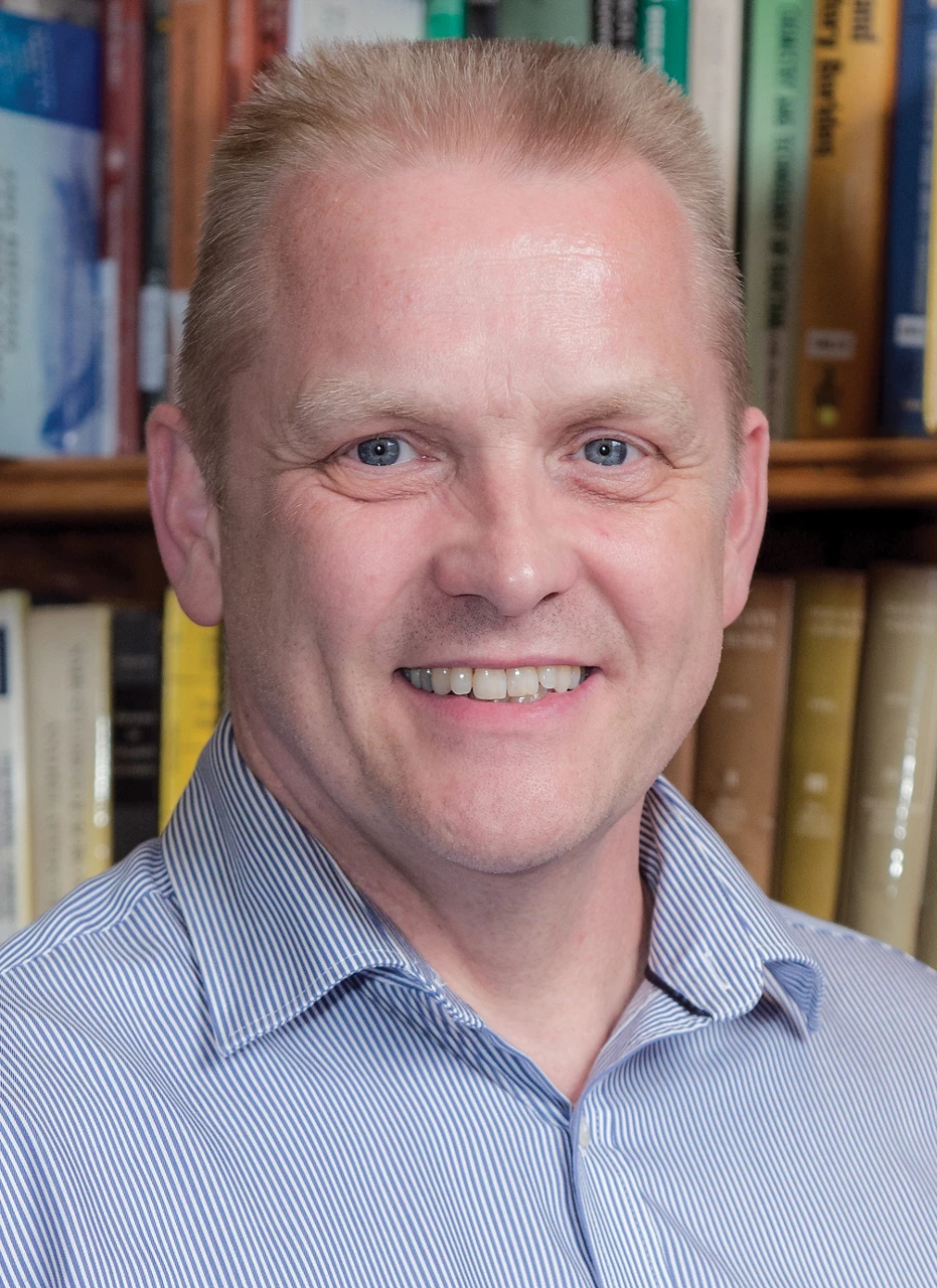 A man looking at the camera, with a bookshelf behind him. He’s wearing a striped shirt and has short hair that stands up.