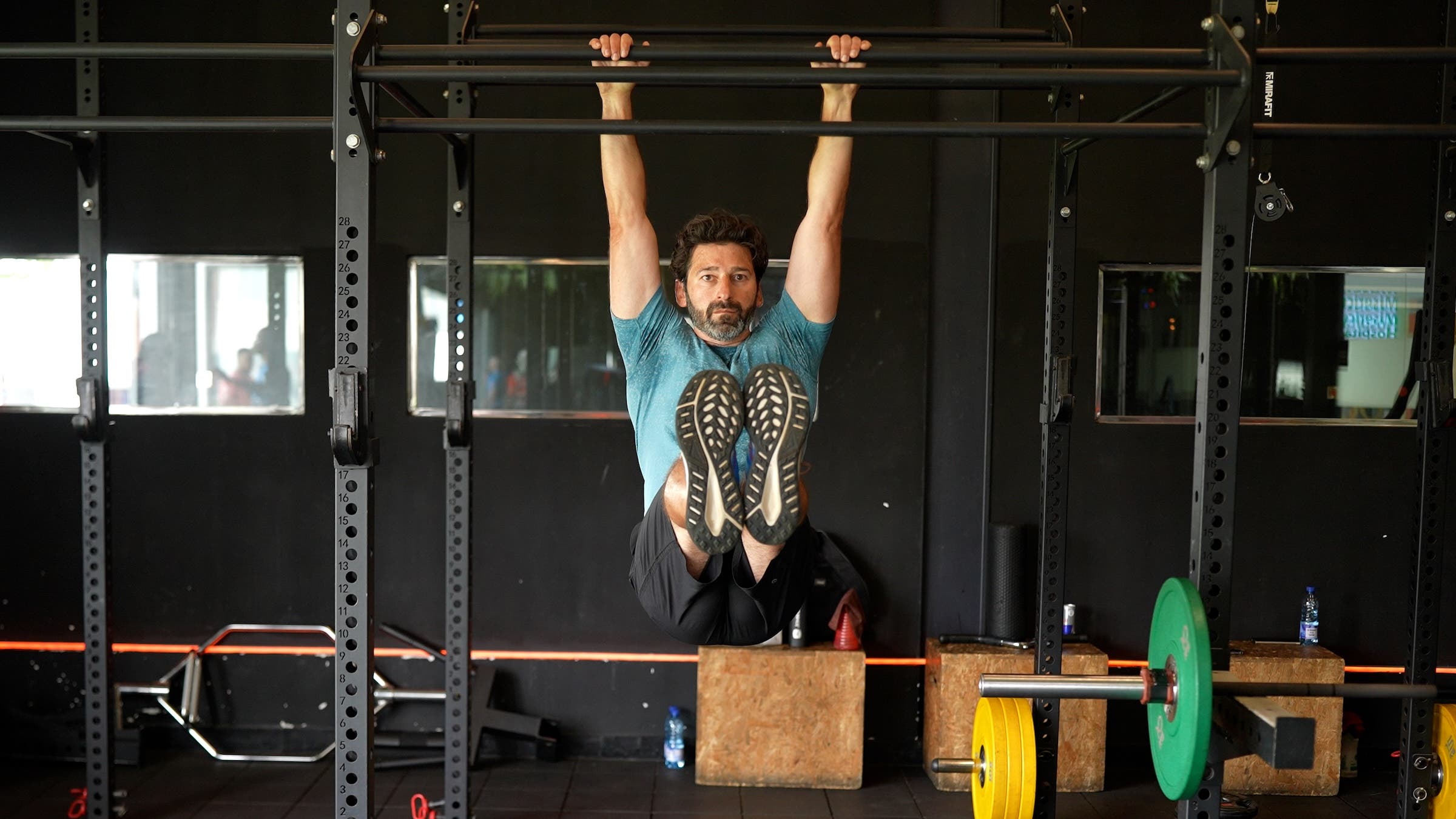 a man with dark hair wearing a blue shirt and black shorts performs a hanging leg raise at a gym