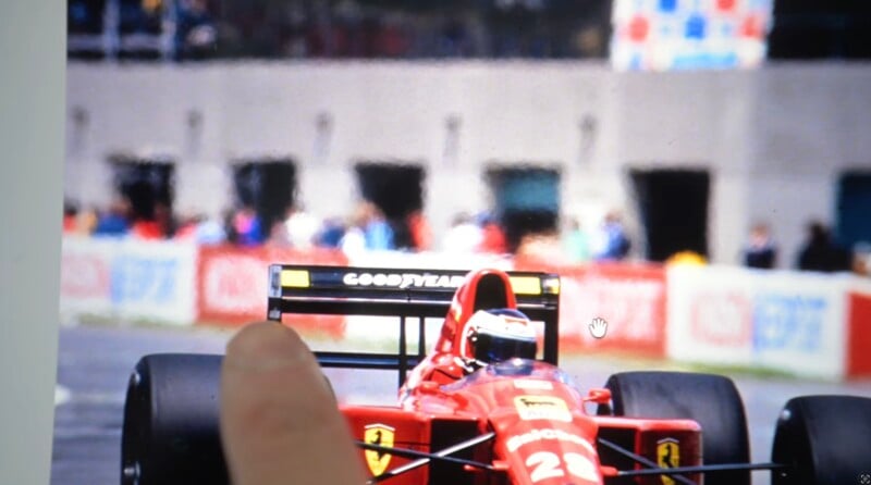 A finger points at the rear wing of a red Ferrari Formula 1 car with number 28 on the nose, on a racetrack with blurred spectators and pit garages in the background.