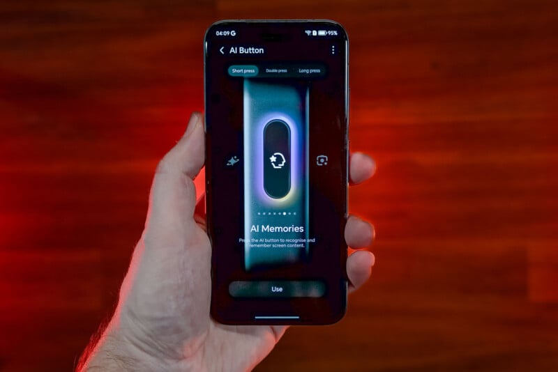 A hand holds a smartphone displaying an “AI Button” screen with options for short and long press. The screen highlights “AI Memories” and has a glowing circular icon in the center. The background is a wooden surface.