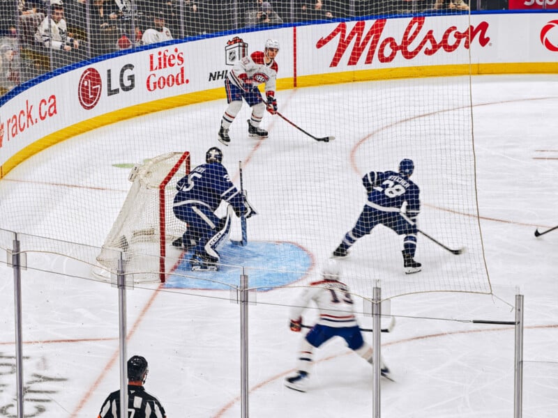 Hockey players from two teams compete near the goal, with a goalie in blue gear ready to defend. Players skate on the ice, surrounded by boards featuring advertisements.