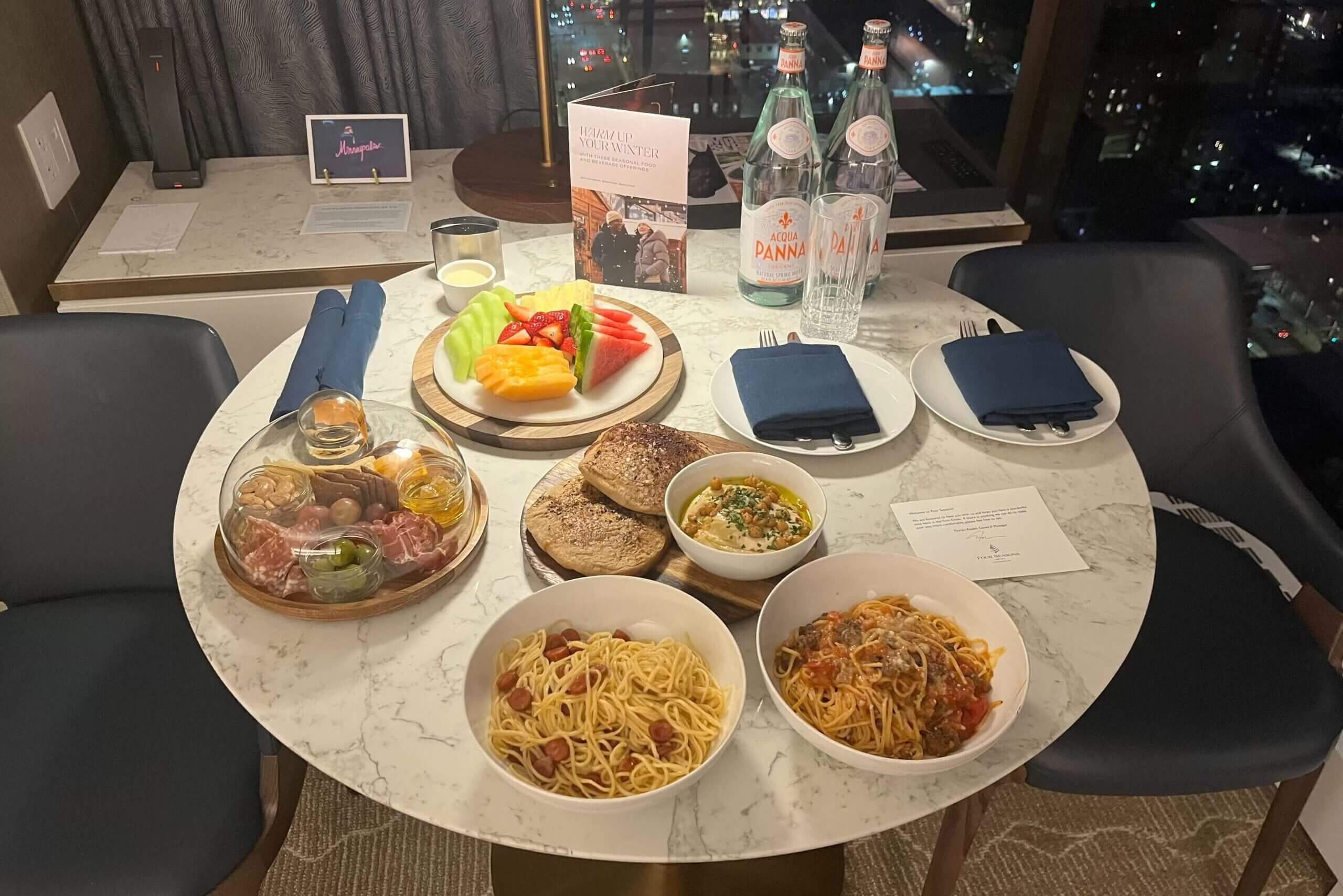 A table with two bowls of pasta, plus a charcuterie board, bread and hummus, and a fruit plate.