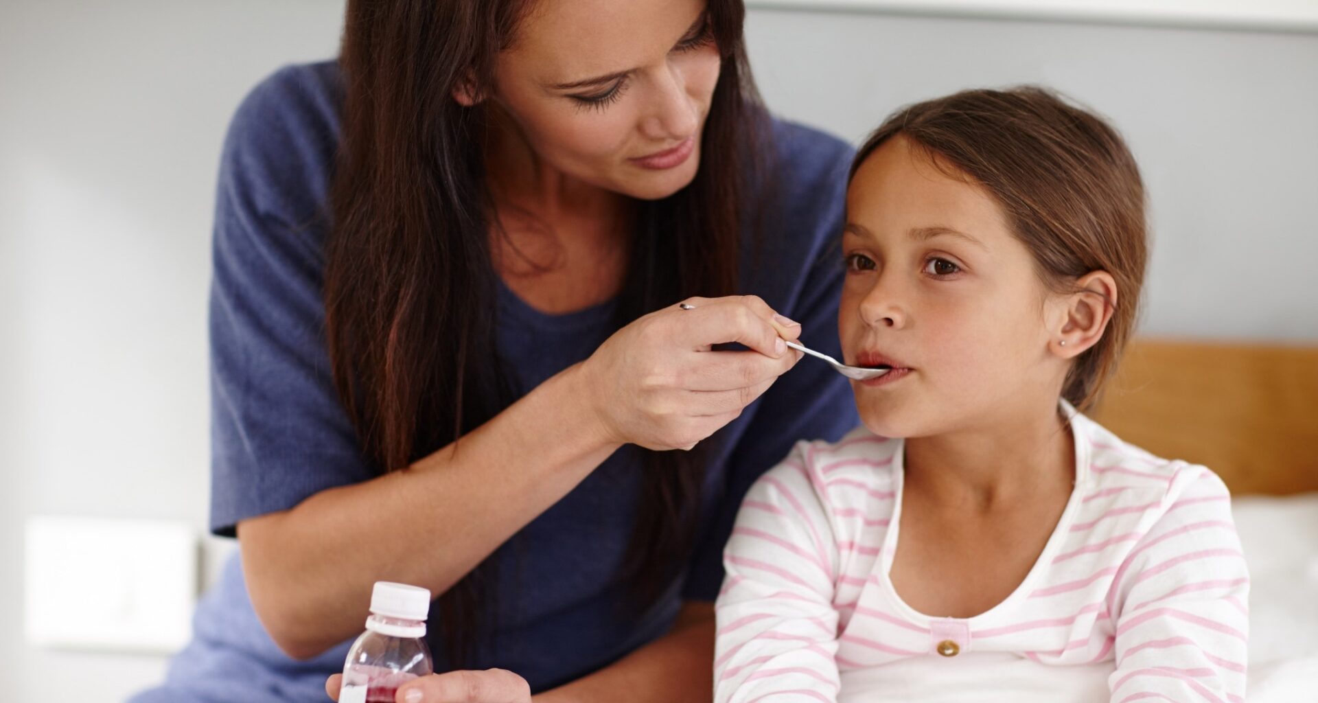 Mother holding a bottle of medicine. Mother giving sick young daughter a spoon of antibiotic medicine. Daughter is in bed.