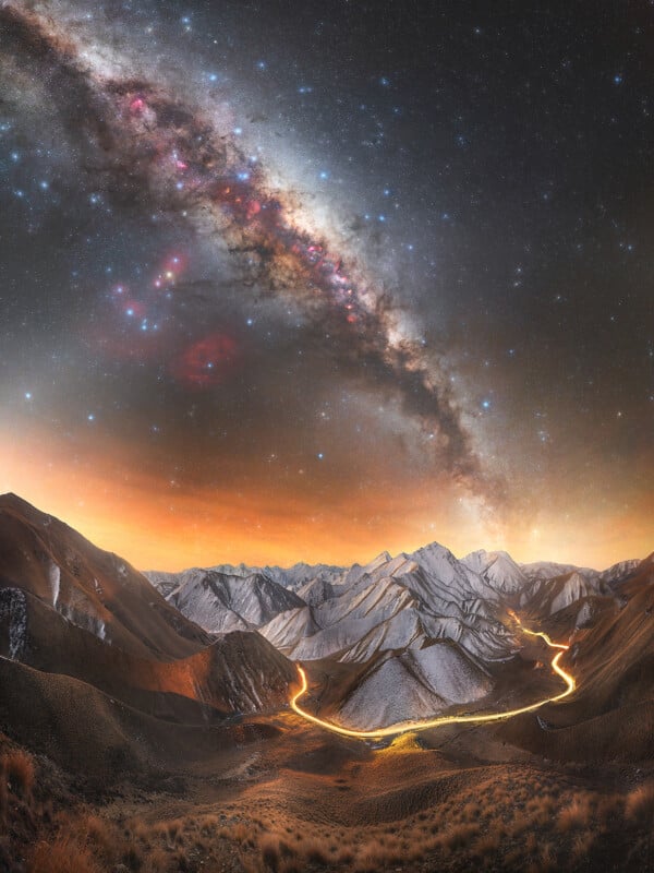 A snowy mountain range under a vibrant night sky with the Milky Way stretching across; a winding, glowing road runs through the mountains, and the horizon is lit with warm, orange light.