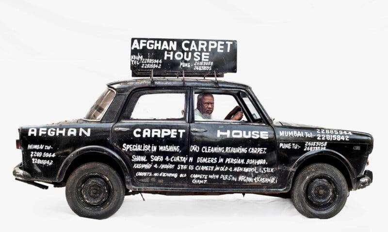 A man sits in a black car painted with white text advertising "Afghan Carpet House" and carpet cleaning services. The car has large signs on the roof and doors listing services and contact details. The background is plain white.