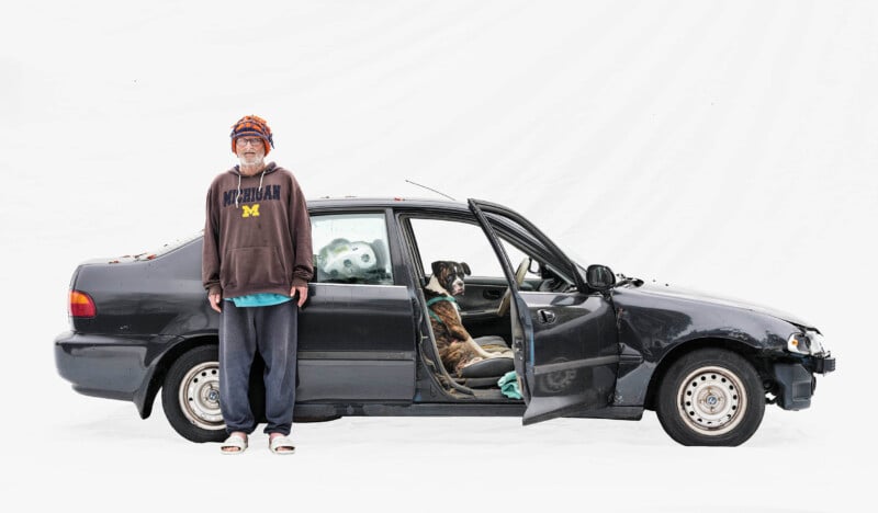 A person in casual clothes and a hat stands beside a black car with the rear door open. Two dogs are inside the car, one sitting in the back seat and the other visible through the window. The background is plain white.