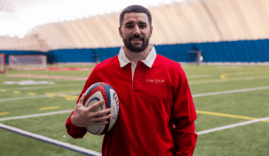 A man with a short beard in a red CSB/SJU rugby shirt stands on an indoor sports field, holding a rugby ball and smiling at the camera. The background shows artificial turf and a domed roof.
