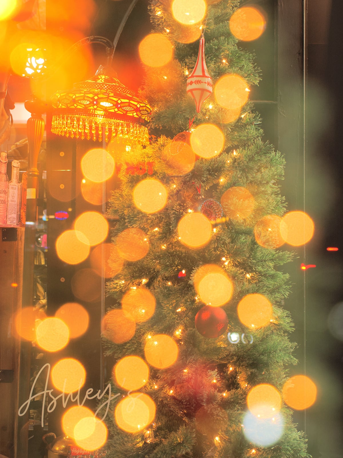 A decorated Christmas tree with ornaments and lights stands indoors, surrounded by warm yellow bokeh lights that create a festive, dreamy atmosphere. A patterned lamp glows nearby.