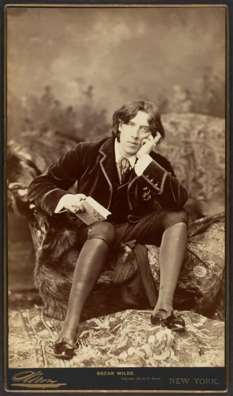 A sepia photograph of a seated man in Victorian attire, resting his head on his hand and holding a book. He sits on a fur-covered chaise with an ornate backdrop, looking thoughtfully toward the camera.