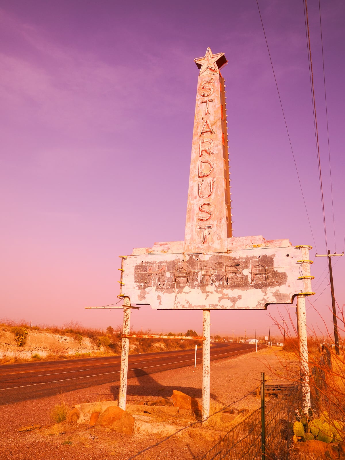 A tall, weathered roadside sign shaped like a star-topped rocket reads "STARDUST." The lower part is faded and illegible. The sign stands by an empty road in a desert landscape under a pinkish sky.