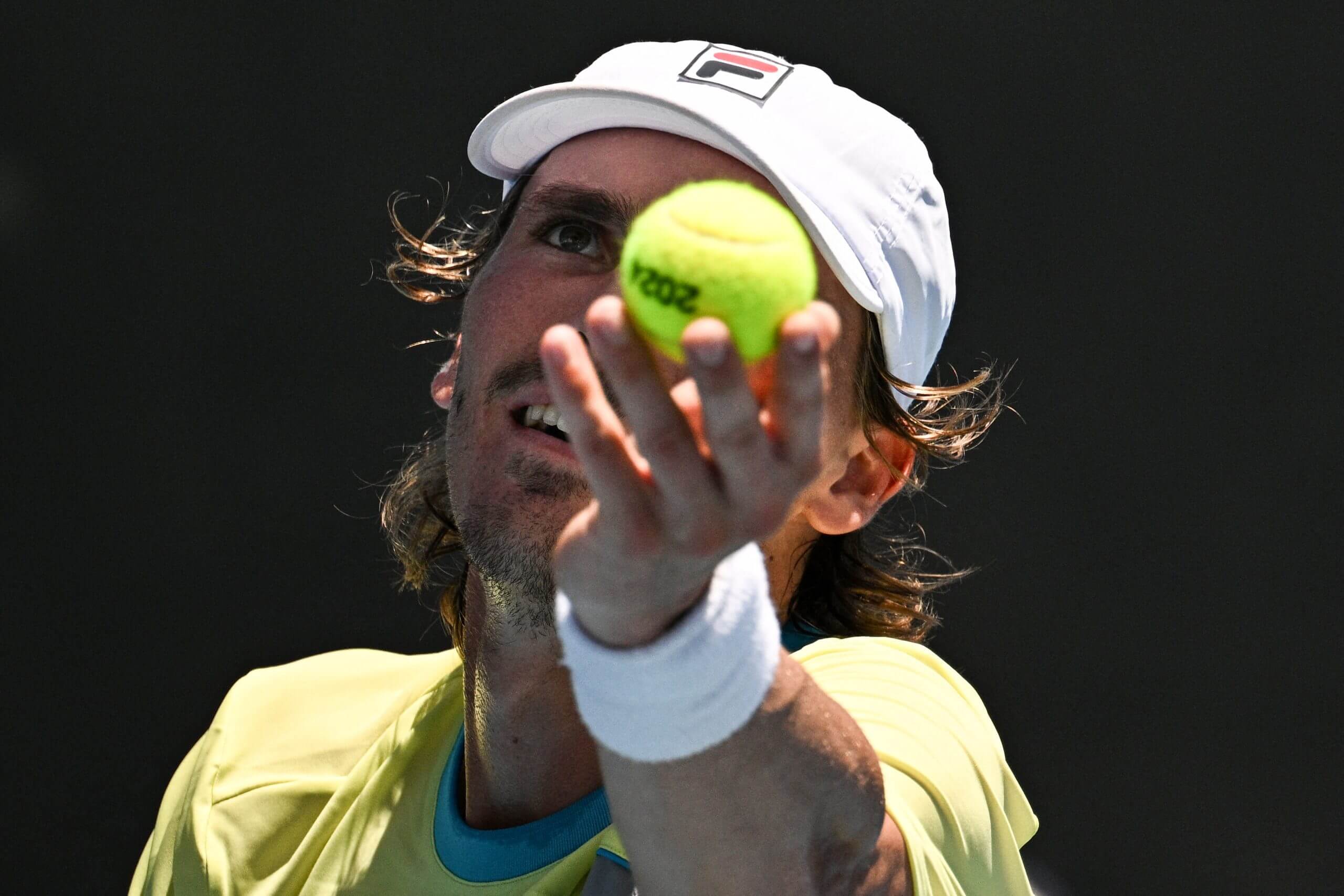 A close-up of Patrick Kypson wearing a white baseball cap and yellow t-shirt as he prepares to toss a tennis ball into the air.