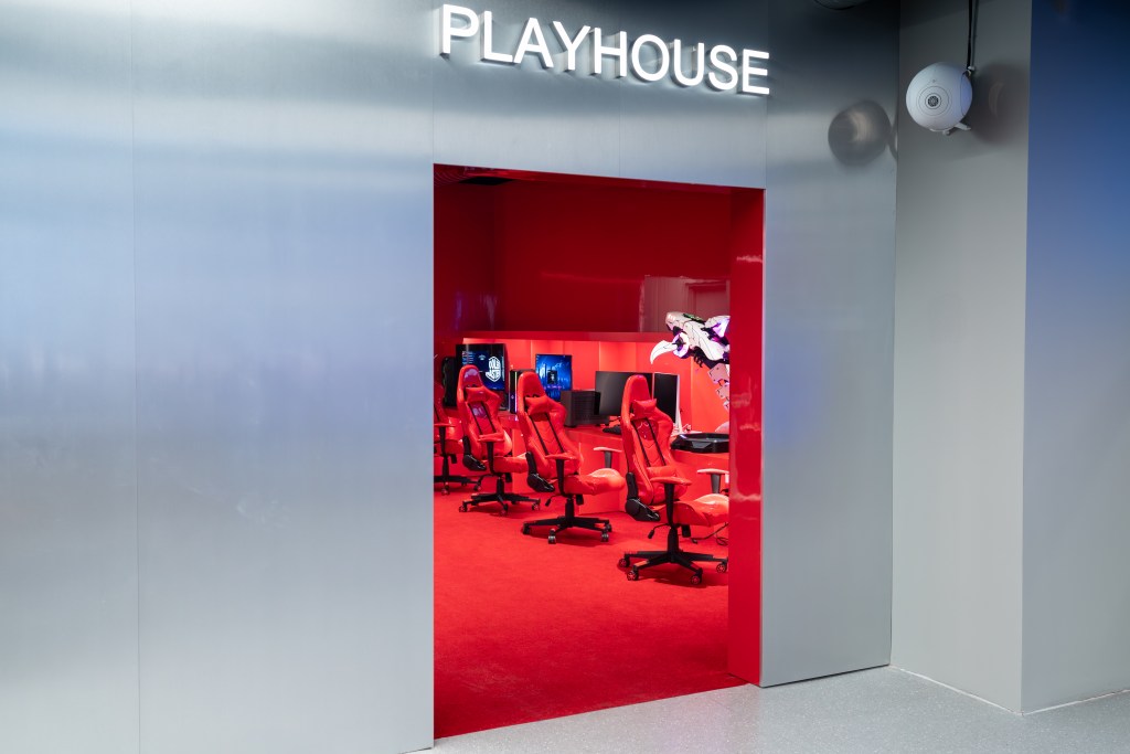 A metallic silver wall with a PLAYHOUSE sign above a doorway, leading into a red room with a row of gaming computers and chairs.
