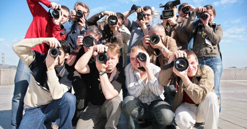 A group of photographers and videographers crouch together outdoors, all aiming their cameras and video recorders directly at the viewer under a bright, clear sky.