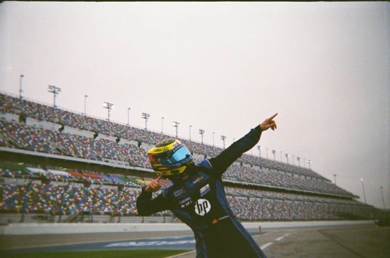 A race car driver in a blue suit and yellow helmet strikes a celebratory pose on the track in front of empty stadium stands at a racetrack.