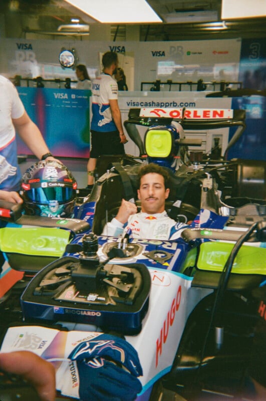 A race car driver sits in a Formula 1 car in a garage, giving a thumbs up. Team members stand nearby, and a helmet rests on the car’s side. The background displays a blue wall with sponsor logos.