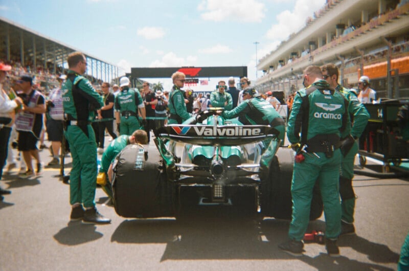 A Formula 1 car with Valvoline and Aramco branding is surrounded by a pit crew in green uniforms on the grid before a race, with grandstands and spectators in the background on a sunny day.