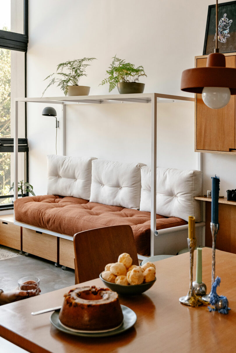 A modern living space with a cushioned daybed, potted plants, a table set with baked goods, candles, and natural light from large windows.