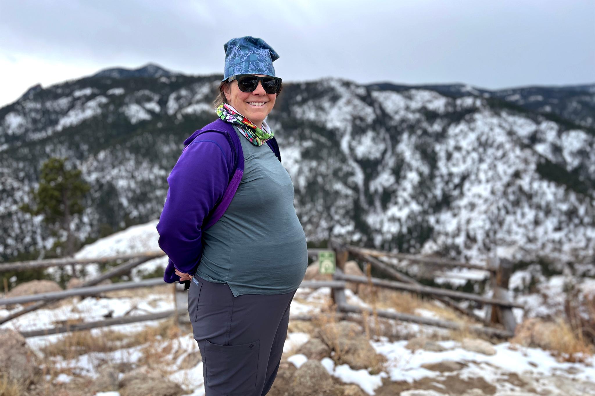 A woman in a purple sweatshirt and shades sticks out her pregnant belly on top of a snow-covered mountain, smiling at the camera.