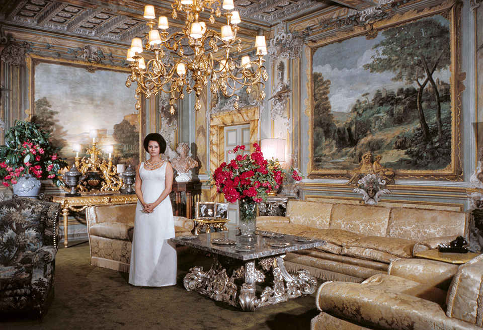 Sophia Loren stands in a long white gown in the center of an opulent, gold-accented drawing room featuring large landscape murals and a crystal chandelier.