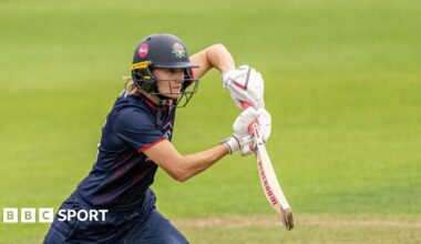 Gaby Lewis playing a shot off the front foot with her bat held high. She is wearing a navy kit with a navy helmet