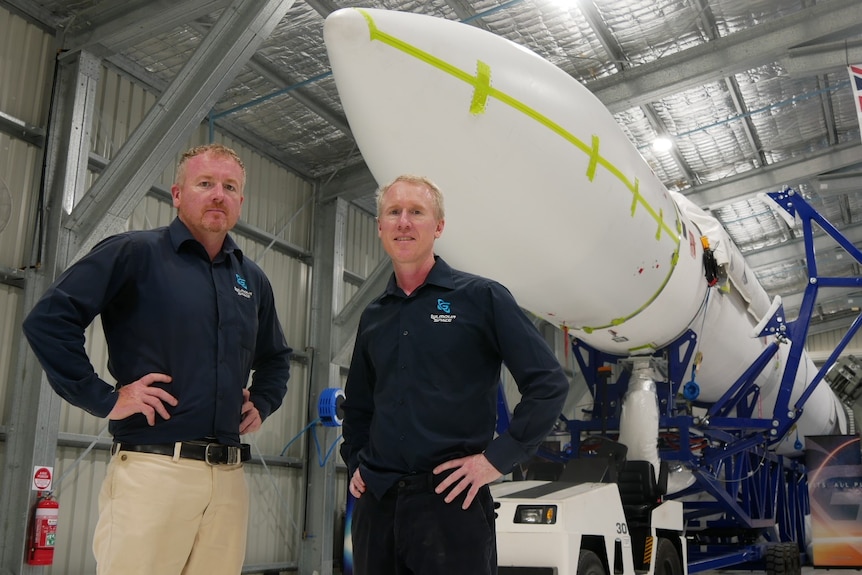Two men stand in front of rocket in warehouse