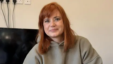 Sally Jones, who is wearing a grey hooded jumper, is sitting on a black leather couch. She is smiling at the camera. 
