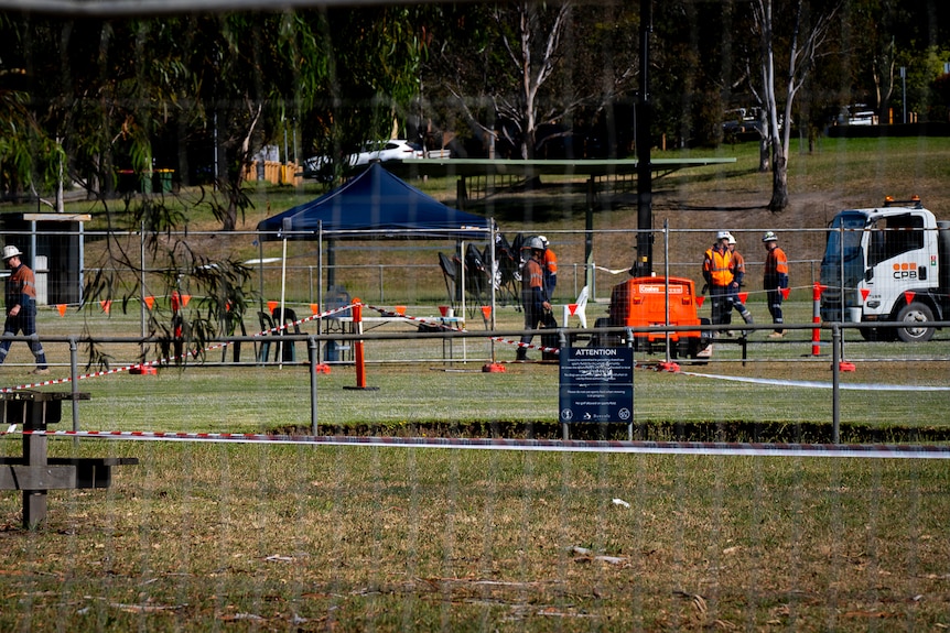 Workers in orange and blue walk behind a fenced off area of an oval where there is a large hole in the ground.