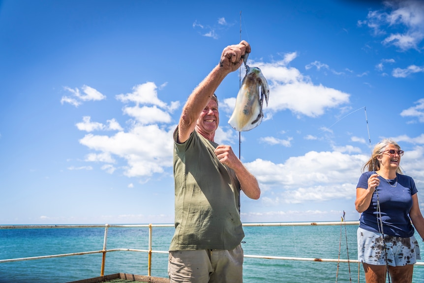 A fisherman pulls his catch off the line on a sunny day, with blue sky, white clouds, and turquoise sea behind him.