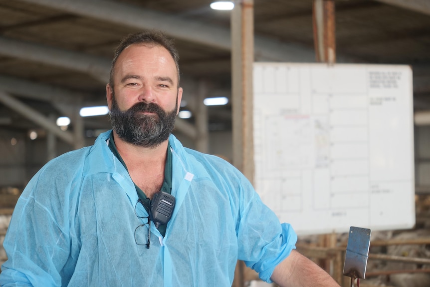 A man with a dark beard stands in front of covered sheep pens.
