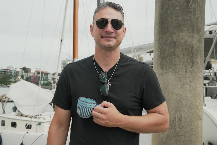 A man wearing sunglasses and a black shirt smiles as he holds a blue mug standing in front of a marina.