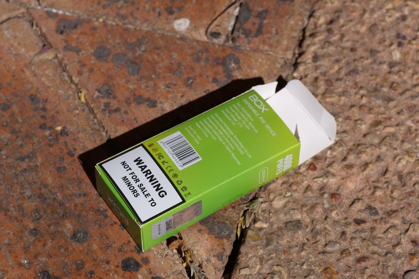 An empty vape box with a label reading "Warning, not for sale to minors" lies on a footpath.